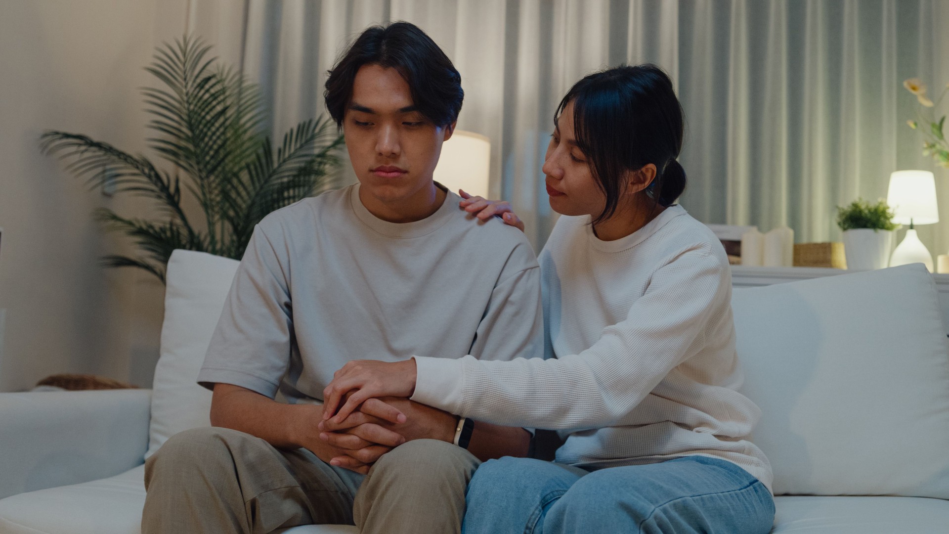 Young Asian woman supporting depressed frustrated boyfriend together on sofa in living room at home night. Lifestyle spend time house.